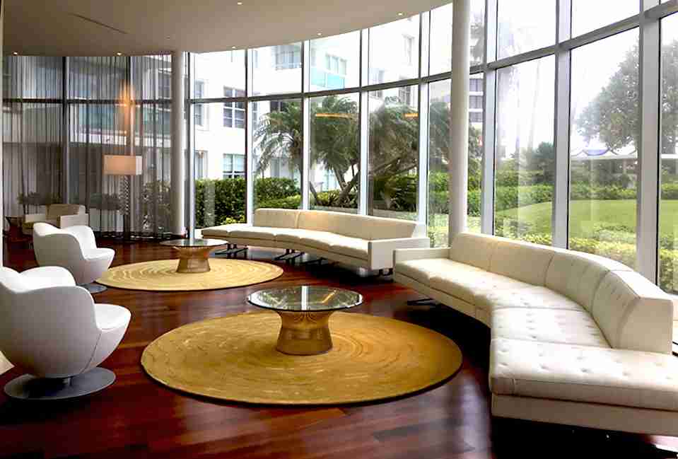 Modern condominium lounge with floor-to-ceiling windows, white curved sofas, round golden rugs, glass coffee tables, and hardwood floors overlooking tropical greenery.