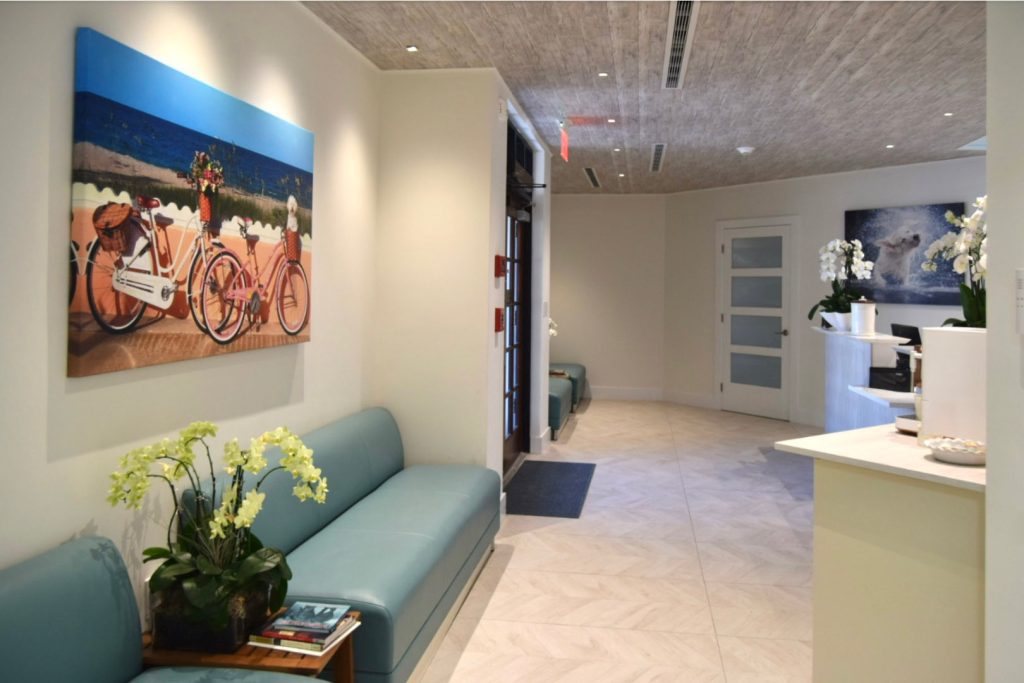 A calm, modern waiting room designed for animals and their owners. The space features comfortable upholstered bench seating, soft neutral walls, and contemporary artwork that adds warmth and color. Light?toned flooring, clean lines, and subtle ceiling textures create an inviting, low?stress environment. Plants and neatly arranged reading materials contribute to a welcoming, well?kept atmosphere near the reception area.